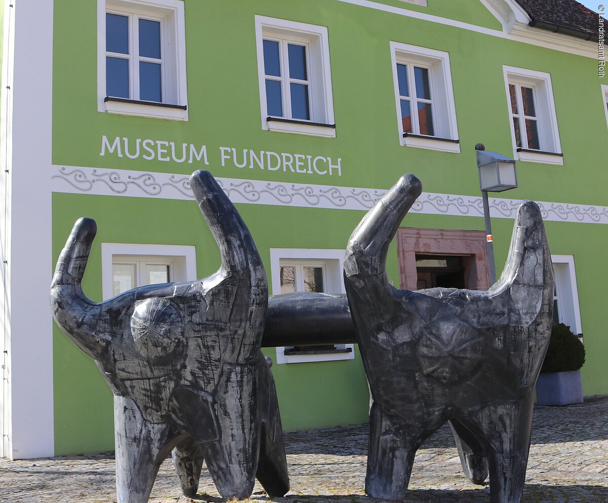 Archäologisches Museum Thalmässing Außenansicht des Archäologischen Museums in Thalmässing. Grünes Haus mit schwarzem Dach. Davor steht eine Skulptur von menschlichen Figuren, die eine Platte tragen.