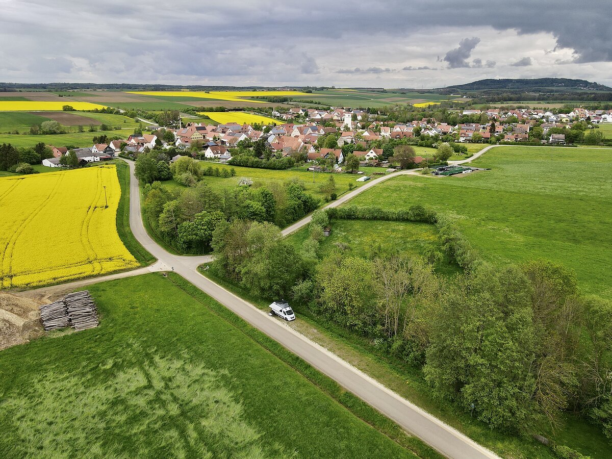 Campingplatz "Märzenbecherwald" Luftaufnahme einer ländlichen Landschaft mit Feldern, einem Dorf und einem Wohnmobil auf einer Straße.