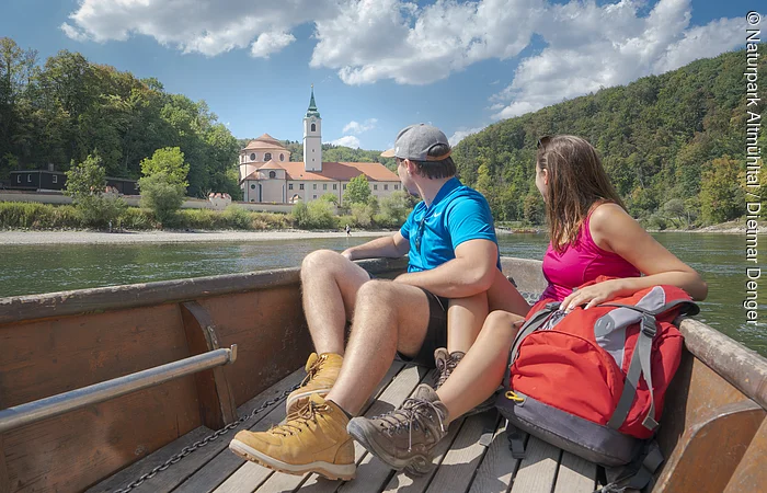 Paar sitzt in einem Boot auf einem Fluss und blickt auf ein Gebäude mit Turm und Wald im Hintergrund.