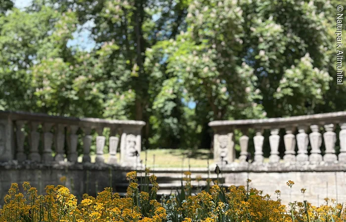 Ein Beet mit Planzen im Vordergrund wird von einer Mauer abgegrenzt. Ein steinerner Weg führt auf eine höhere Ebene mit Bäumen