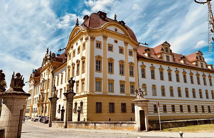 Zentrales Element des Bildes ist die Schlossresidenz Ellingen mit kräftigen Farben vor einem blauen Himmel. Links ist ein Pfeiler mit einer Statue zu sehen
