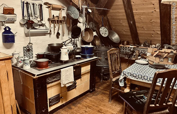 ein alter Holzherd mit Kochutensilien und ein gedeckter Tisch mit Stühlen ist zu sehen.