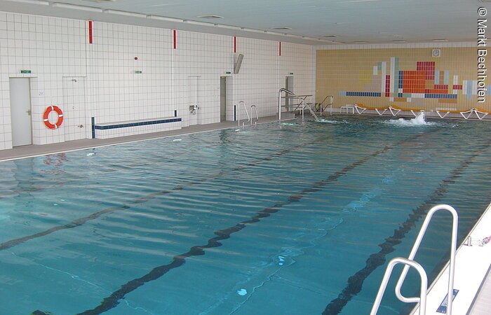 Ein rechteckiges Schwimmbecken des Hallenbads in Bechhofen. Das Schwimmbecken nimmt den gesamten Raum ein. Die Wände im Hintergund sind bunt und weiß gekachelt.