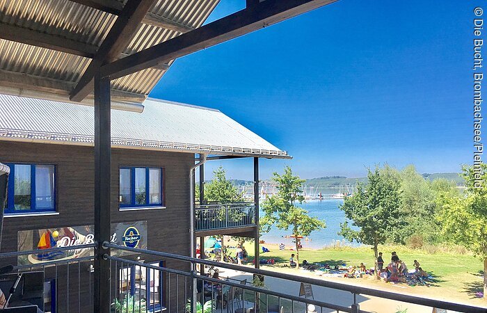 Blick von einem Balkon auf ein zweistöckiges Holzhaus mit Terasse. Dahinter eine Liegewiese mit See.