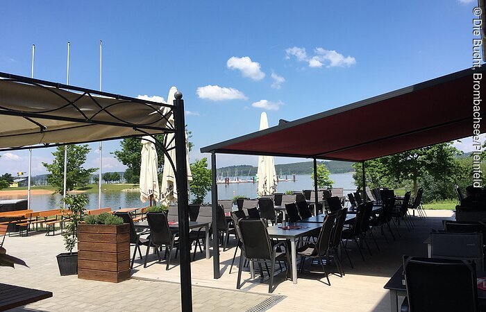 Eine große geplasterte Terasse am See. Im Vordergrund eine Markisse und ein Pavillon. Schwarze Stühle und Tische stehen in der Sonne.