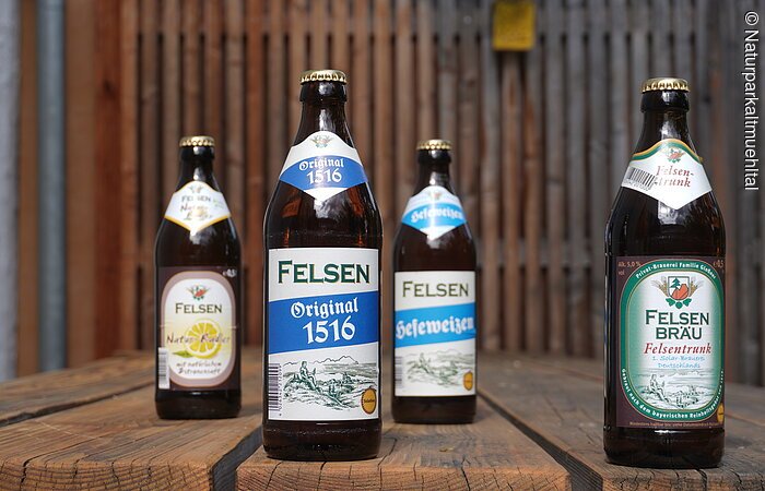 Vier Bierflaschen der Marke Felsen stehen auf einem Holztisch vor einem Holzzaun.