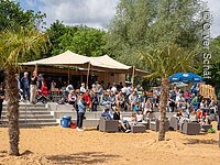 Wakeboardanlage Eine Tribüne aus Stein dirket an einem Sandstrand. Darauf sitzen viele Menschen. Dahinter ein Sonnensegel und grüne Bäume.