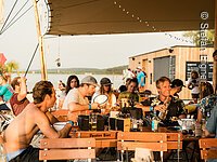 Wakeboardanlage Gäste sitzen unter einem Sonnensegel an Tischen aus Holz. Im Hintergrund ein Wakeboard-Verleih direkt am See.