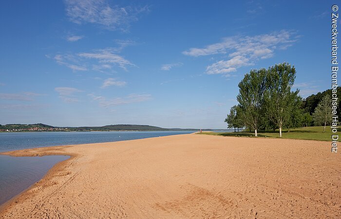 Breiter, flacher Sandstrand in Allmannsdorf am großen Brombachsee. Im Hntergund beginnt links der Brombachsee und rechts eine Liegewiese mit Bäumen.