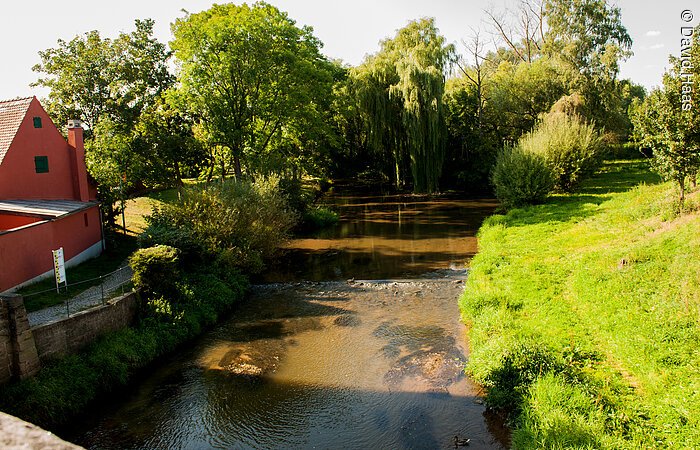 Blick von einer Brücke auf einen Fluss. Links ein Haus mit roter Fassade. Am Ufer wachsen Bäume. Am rechten Uferrand eine grüne Wiese.