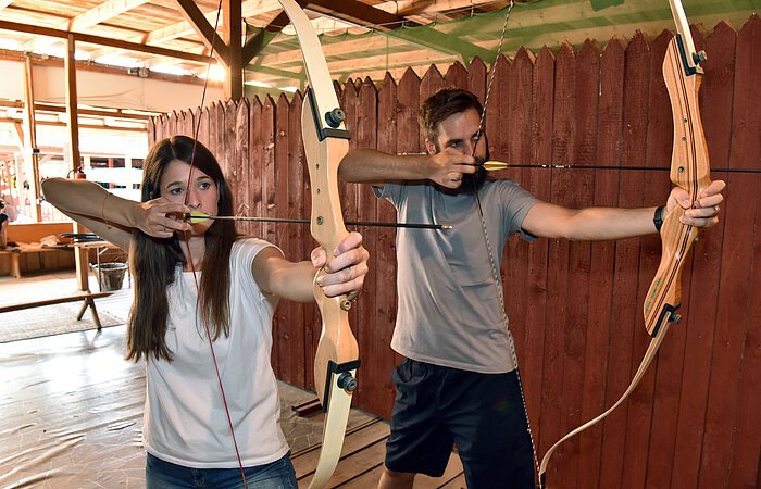 Eine Frau und ein Mann halten jeweils einen gespannten Bogen und zielen mit dem Pfeil. Im HIntergudn ein hoher Holzzaun.