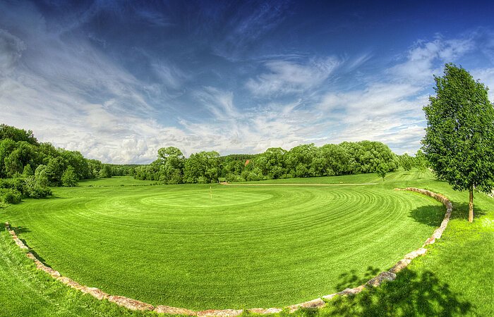 Große grüne Wiese eines Golfplatzes. Der PLatz ist umgebennvon Bäumen und Sträuchern.