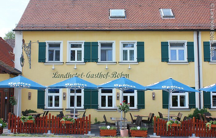 Teilansicht eines zweistöckigen Hauses im fränkischen Baustil mit gelber Fassade und grünen Fensterläden. Davor ein kleiner Biergarten mit braunem Zaun und blauen Sonnenschirmen.