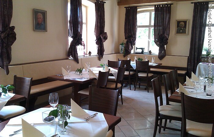 Blick in einen Gastraum mit beigen Wänden und braunen Vorhängen. Entlang der Wände Bänke. Davor Tische und Stühle aus dunkelbraunem Holz. Die Tische sind festlich mit weißen Tischdecken und Servietten gedeckt.