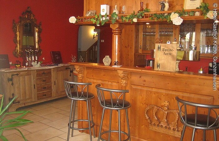 Eine Bar aus Holz mit drei Barhockern. In den Regalen hinter der Bar stehen Gläser. Links von der Bar im Hintergund ein Sideboard aus hellem Holz vor einer rot gestrichenen Wand.