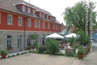 Außensicht auf das Hotel- Gasthof Krone in Gunzenhausen mit Biergarten und Parkplätzen. Das Gebäude ist rot gestrichen. Das Erdegschoss ist gemauert. Auf der Terasse rechtsv neben dem Gbäude steht ein großer grüner Baum und weiße Sonnenschirme.