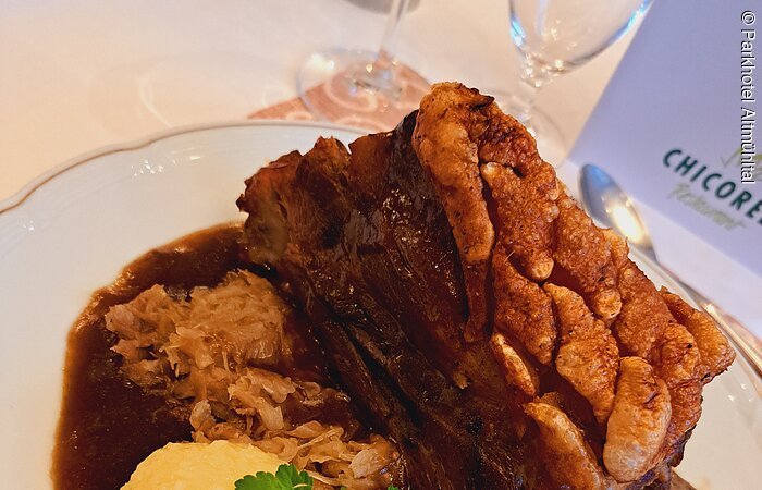 Zwei Kartoffelknödel mit Petersilie garniert liegen auf Bratensoße. Dahinter ein schönes, knuspriges Stück Schäuferle. Ein Sektglas ist zur Hälfte zu sehen.