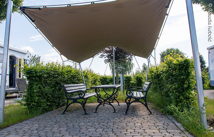 Zwei Bänke und ein Tisch aus Holz stehen unter einem Sonnensegel umgeben von grünen Sträuchern. Im Hintergund erkennt man stehende Wohnmobile.