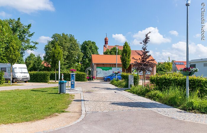 Am Ende eines geplasterten Weges steht ein kleines Häuschen unter blauem Himmel. Gegenüber steht ein weißes Wohnmobil. Links und rechts des Weges sind grüne Sträucher und Wiese.