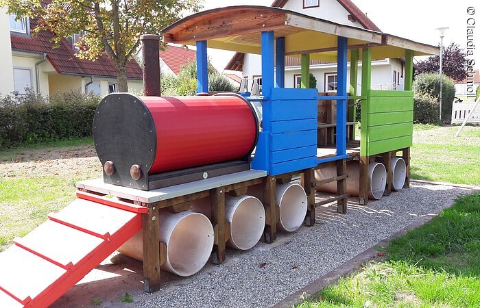 Ein großes Spigerät aus bunt lackiertem Holz. Das Spielgerät hat die Form einer Eisenbahnlok.