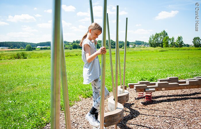 Ein junges Mädchen balanciert über einen Balken in Schlangenform. Dabei hält sie sich an Metallstangen fest. Im Hintergund grüne Wiesen.