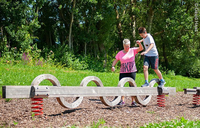 Ein Junge balanciert über einen langen Holzbalken der auf zwei Sprungfedern befestigt ist. Eine Frau hält ihn an der Hand und stützt ihm beim Laufen über den Balken. Im Hintergund Wald und Wiese.