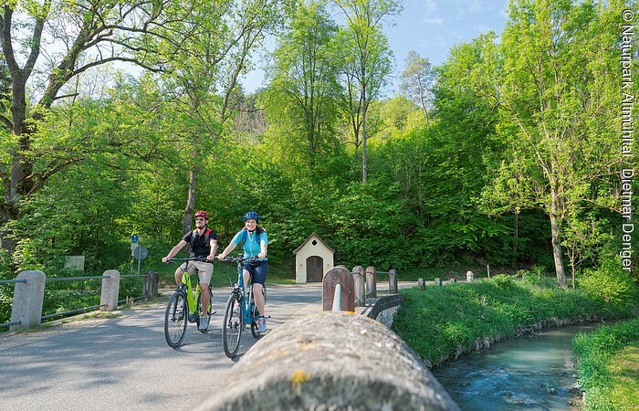 Ein Pärchen ist mit ihren Fahrrädern auf dem Radweg unterwegs. Rechts begleitet sie die Anlauter. Der Wald strahlt in hellem grün. Hinter ihnen sieht man eine kleine Kapelle.