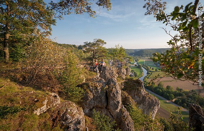Ein Wanderpärchen, etwas entfernt, steht links auf einem Felsen an der Arnsberger Leite, die gut zu sehen ist. Dahinter erstreckt sich der Blick ins Tal. Der Himmel erstrahlt wolkenlos.