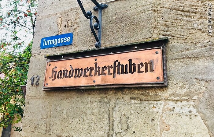Handwerkerstuben Ein Schild aus Kupfer. Drauf steht Handwerkerstuben. Das Schild hängt an einer Steinmauer. Darüber ein kleines blaues Straßenschild mit der Aufschrift Turmstraße.