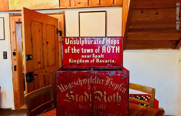 Hopfenversendungsbox Eine rote Kiste aus Metall steht auf eibnem Tisch in einem Wohnraum. Auf der Kiste steht in weißer Schrift: Unsulphurated Hops of the town of Roth near Spalt Kingdom of Bavaria