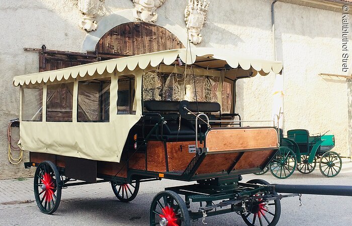 Eine große Kutsche aus Holz steht in einem Hof. Die Kutsche hat eine Zeltüberdachung.