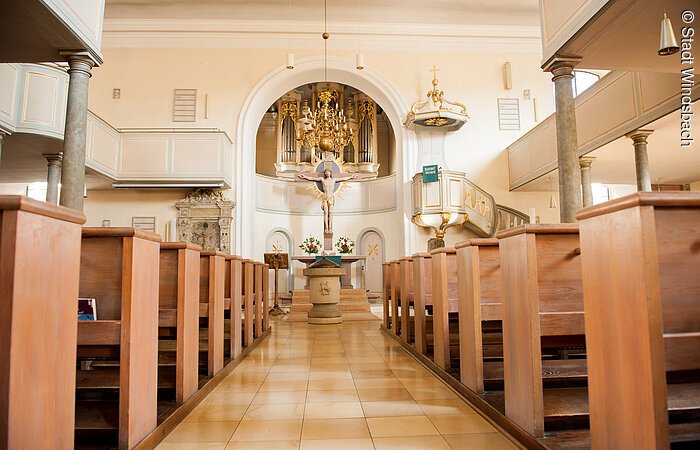 Innenraum einer Kirche. Links und rechts zwei Emporen. Darunter Holzbänke. In der Mitte steht ein Holzkreuz mit Jesus Christus. Darüber hängt ein goldener Kronleuchter von der Decke.