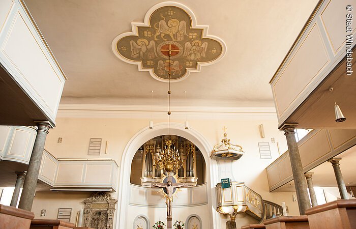 Innenraum einer Kirche. Links und rechts zwei Emporen. In der Mitte steht ein Holzkreuz mit Jesus Christus. An der weißen Decke ein braunes Relief. Mittig davon hängt ein Kronleuchter von der Decke.