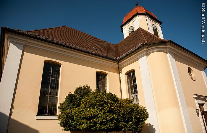 Blick von unten auf die Stadtkirche Windsbach. Eine Kirche mit weiß-gelber Fassade. Hinter dem Kirchendach spitzt der Kirchturm hervor.
