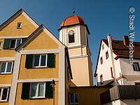 Blick von unten auf eine Kirchturmspitze. Ein Turm mit weiß-gelber Fassade und rotem Haubendach. Davor stehen Häuser im fränkischen Baustil. Links ein gelbes Haus mit grünen Fensterläden. Rechts ein Haus mit weißer Fassade.