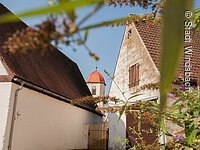 Ausschnitt von zwei älteren Häusern. Zwischen den Häusern ragt ein Kirchturm hervor. Der Turm hat eine weiß-gelbe Fassade mit rotem Haubendach.