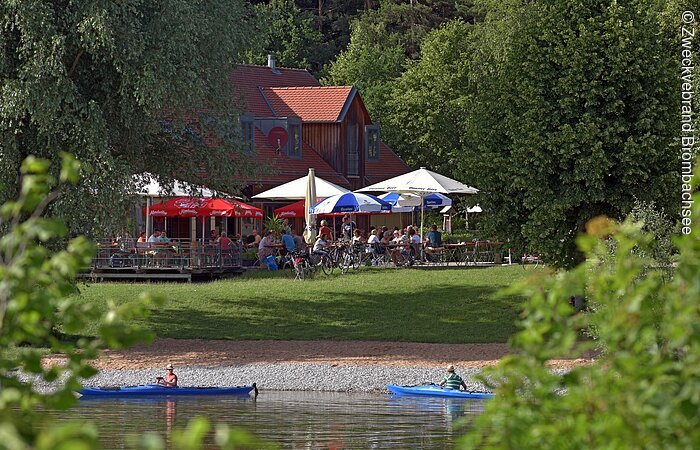 Blick auf den See mit Kanufahrern und den Kiosk Seeklause Absberg mit Sitzmöglichkeiten und Sonnenschirmen am Ufer.
