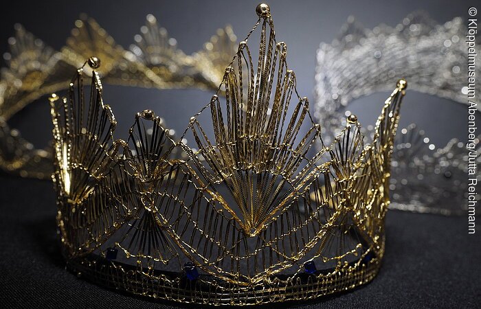 geklöppelte Krone Eine geklöppelte goldenen Krone liegt auf einem grauen Untergrund.