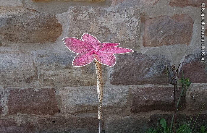 geklöppeltes Mauerblümchen Eine geklöppelte rosa Blume steht in einer Grünfläche vor einer Mauer.