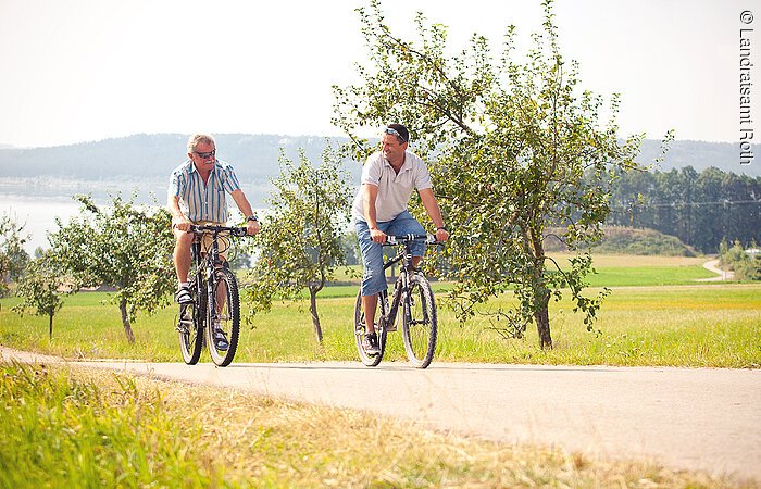 Zwei Radfahrer fahren auf einem Fahrradweg entlang grüner Weiesen. Am Wegesrand stehen Obstbäume.