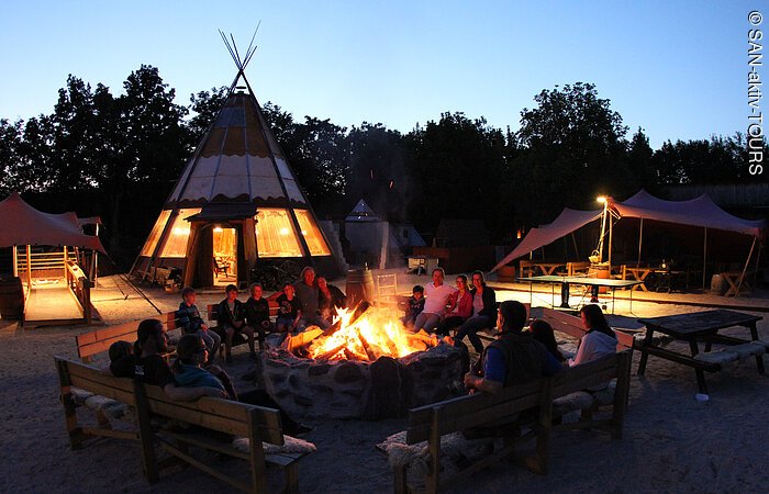 Ein Gruppe Menschen sitzt in der Abenddämmerung auf Holzbänken um ein großes Lagerfeuer. Im Hintergrund stehen ein Tippi und ein Sonnensegel.