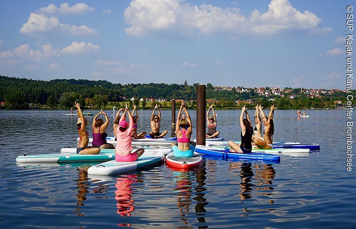 Eine Gruppe von Sportlern sitzt auf ihren Standuppaddle in einem Kreis. Die Bretter schwimmen auf dem See. Alle Sportler machen die gleich Yogapose und strecken ihre Hände in die Luft.