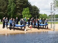 Zwei große Gruppen an Sportlern halten gemeinschaftlich jeweils ein großes Surfbrett. Die Bretter sind in etwa 2 Meter lang. Die Sportler tragen Neoprenanzüge und stehen an einem Strand mit Paddeln in der Hand.
