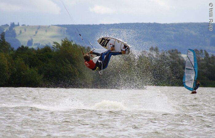 Ein Kitesurfer, der mit seinem Brett in der Luft hängt. Im Hintergund ein Windsurfer, der über den See fährt.