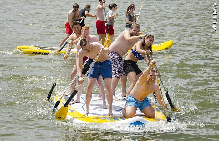 Eine gruppe in Badekleidung steht auf einem großen Stand- Up Paddlingbrett und padelt durch das Wasser. Das Brett ist in etwas zwei mal drei Meter groß. Auf dem Brett stehen 6 junge Erwachsene. Dahinter eine andere Gruppe die über das Wasser paddelt.