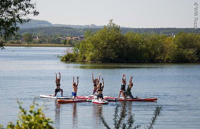 Eine Gruppe von sehcs Sportlerin macht auf ihren Surfbrettern Yoga auf dem See. Die Bretter sind in einem Kreis angeordnet.