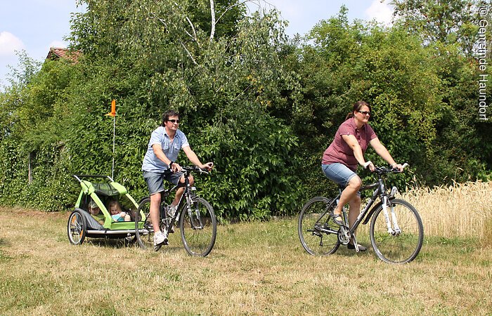 Eine Frau und ein Mann mit Fahrradanhänger radeln über eine Wiese. Im Anhänger sitzen zwei kleine Kinder.