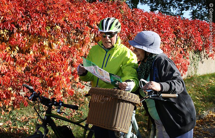 Zwei Radler stehen vor einer mit roten Blättern bewachsenen Mauer und schauen in eine Übersichtskarte.