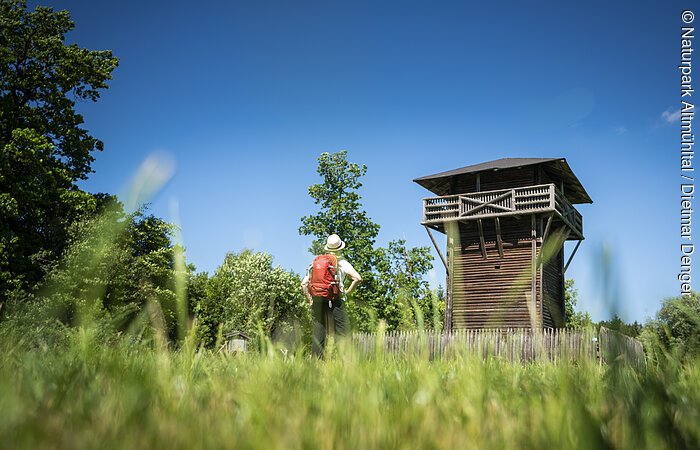 Wanderer mit rotem Rucksack steht auf Wiese vor hölzernem Aussichtsturm bei blauem Himmel