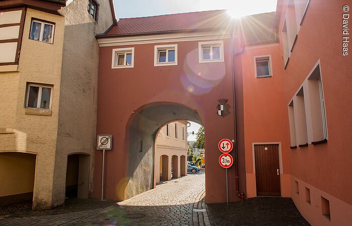 Stadttor Windsbach Ein Stadtor eingebunden zwischen zwei Häusern. Durch das Tor führt eine gepflasterte Straße der Altstadt. Über dem Torbogen drei kleine Fenster, dann folgt das Dach. Das Tor hat eine rötliche Fassade. Die Sonne spitzt über den Dächern hervor.